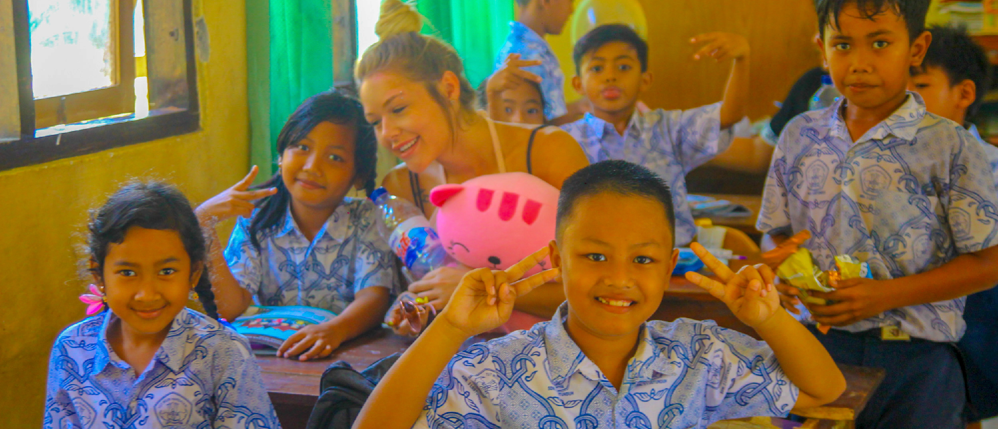 Balinese schoolchildren and a volunteer smiling and making peace signs in a bright classroom.