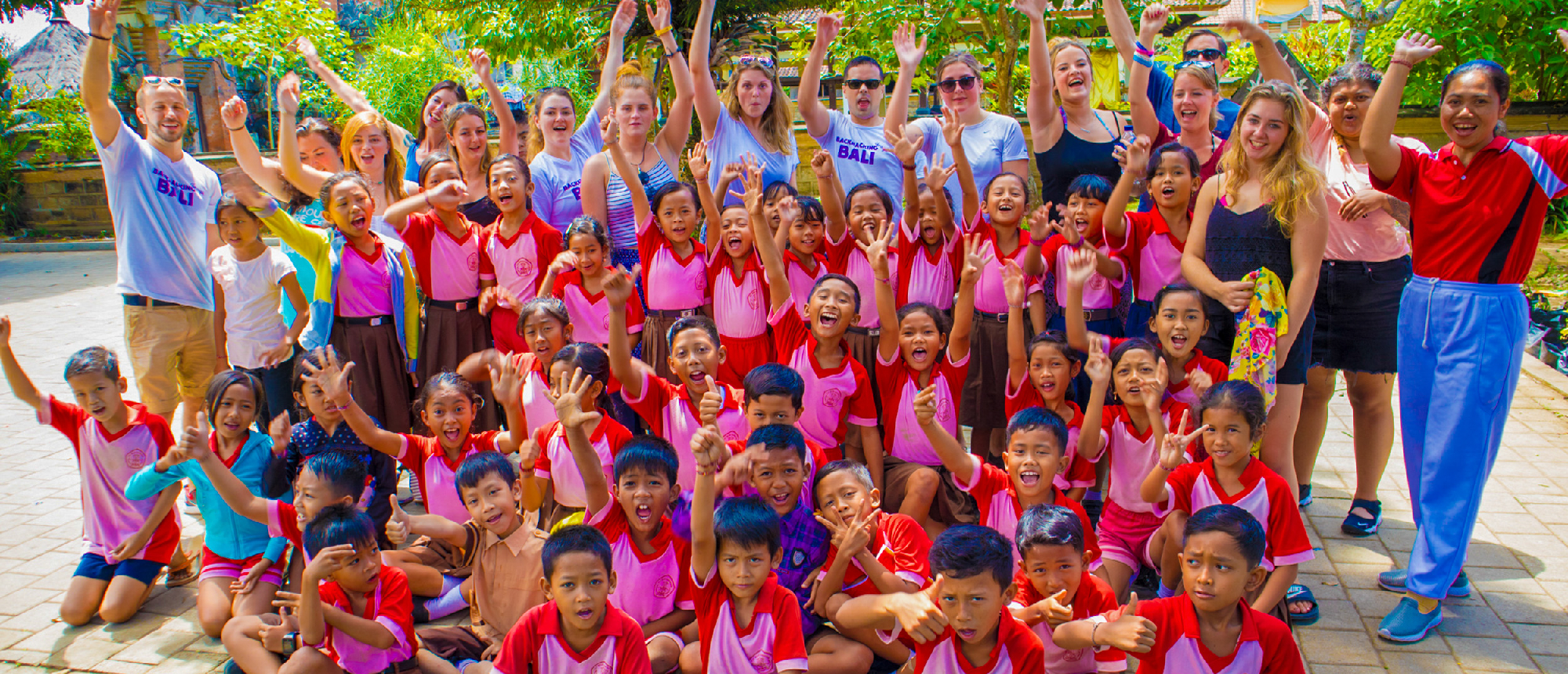 A large group of smiling children and adults enthusiastically wave their hands at a school in Ubud, Bali.