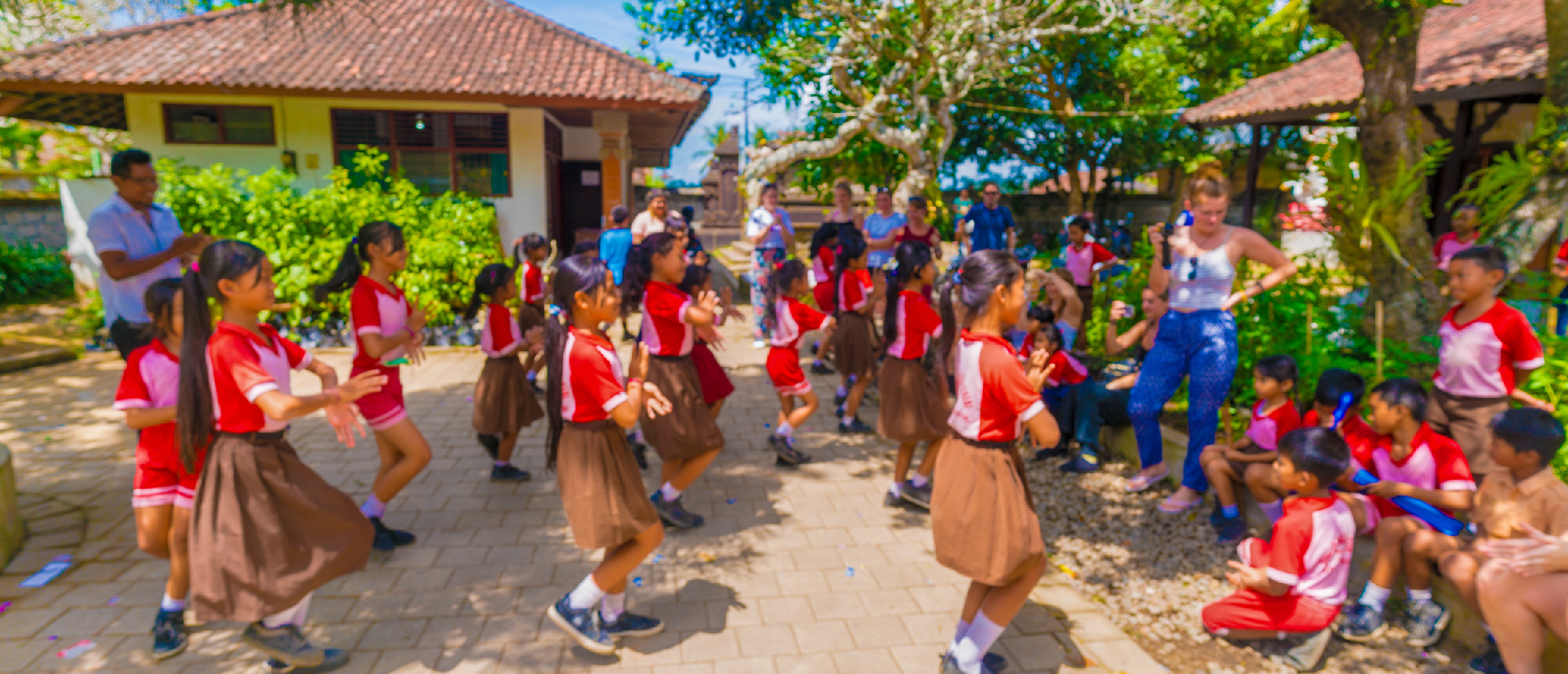 Smiling Balinese students in school uniforms joyfully dance outdoors in a sunny Ubud schoolyard.