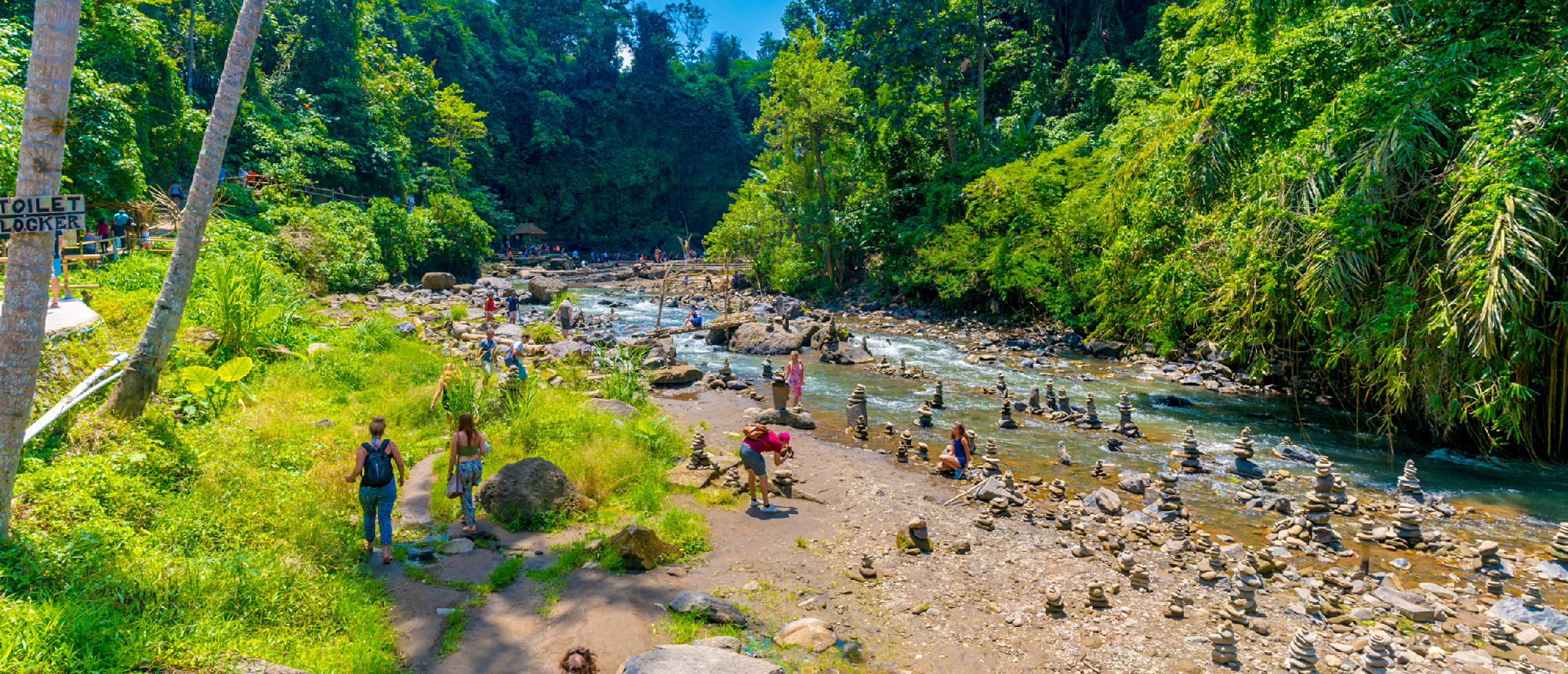 Travelers explore a rocky riverbed with stacked stones amidst lush green jungle in Ubud, Bali.