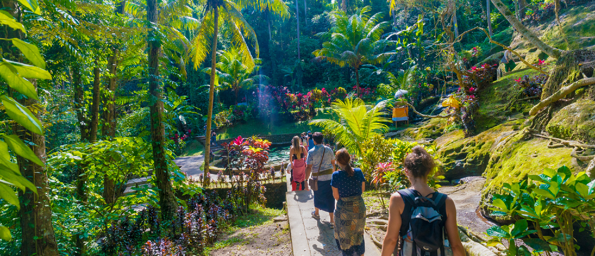 Backpackers walk a lush tropical path surrounded by vibrant greenery and colorful flowers in Bali.