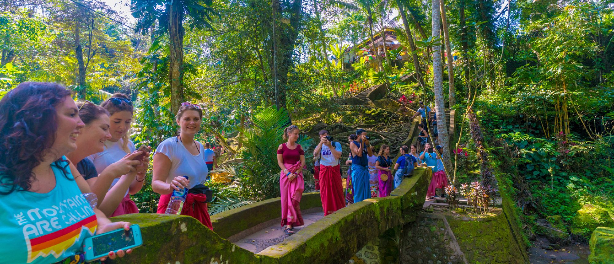 A group of happy backpackers walk along a mossy stone path in a lush jungle in Ubud, Bali.