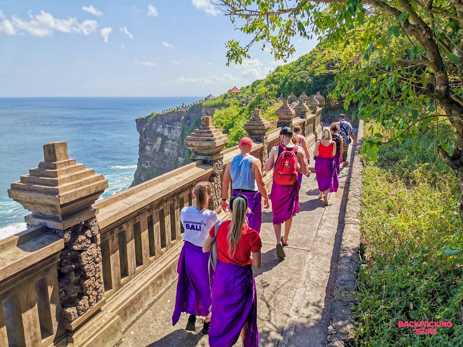 Backpackers in traditional sarongs walk a scenic cliffside path overlooking the ocean at Uluwatu, Bali.