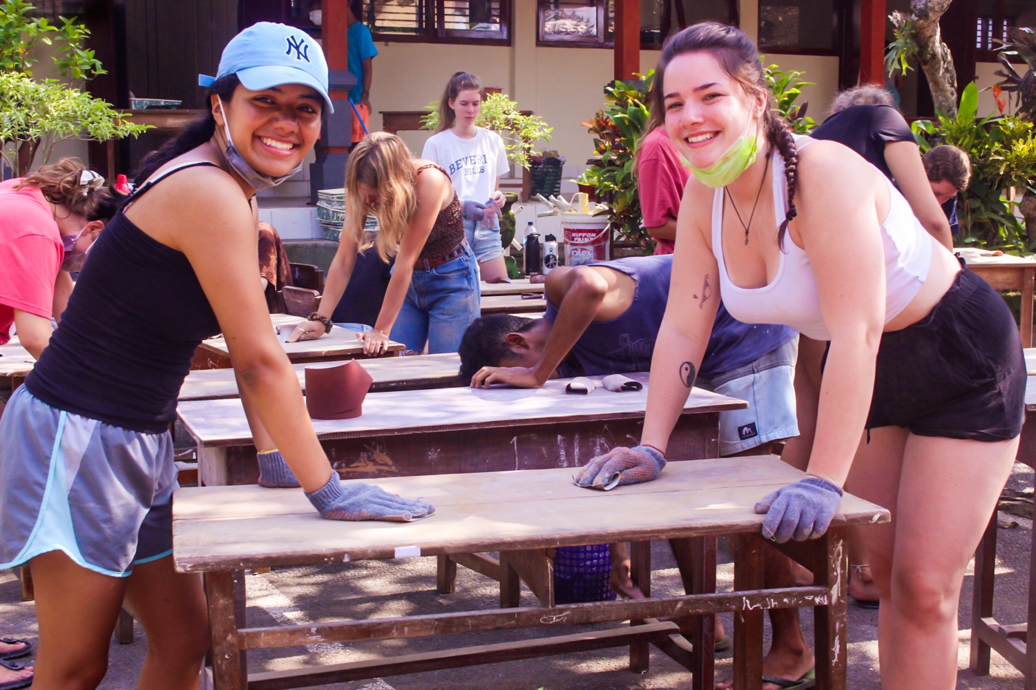 Two smiling women sanding wooden desks outdoors during a community renovation project in Bali.