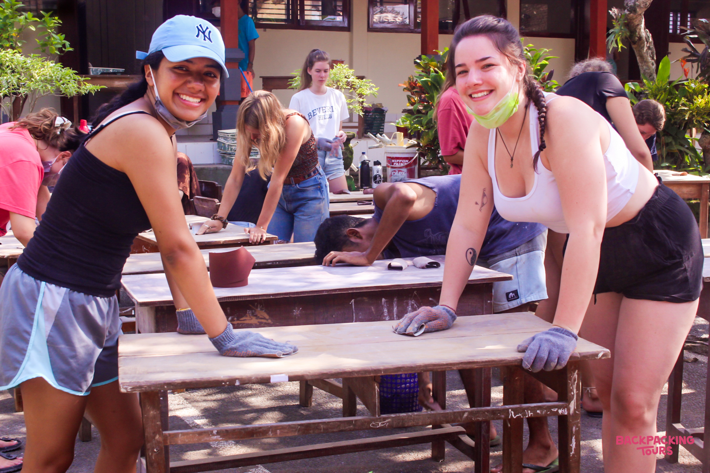 Smiling women in gloves sanding old wooden desks during a community renovation project in Bali.