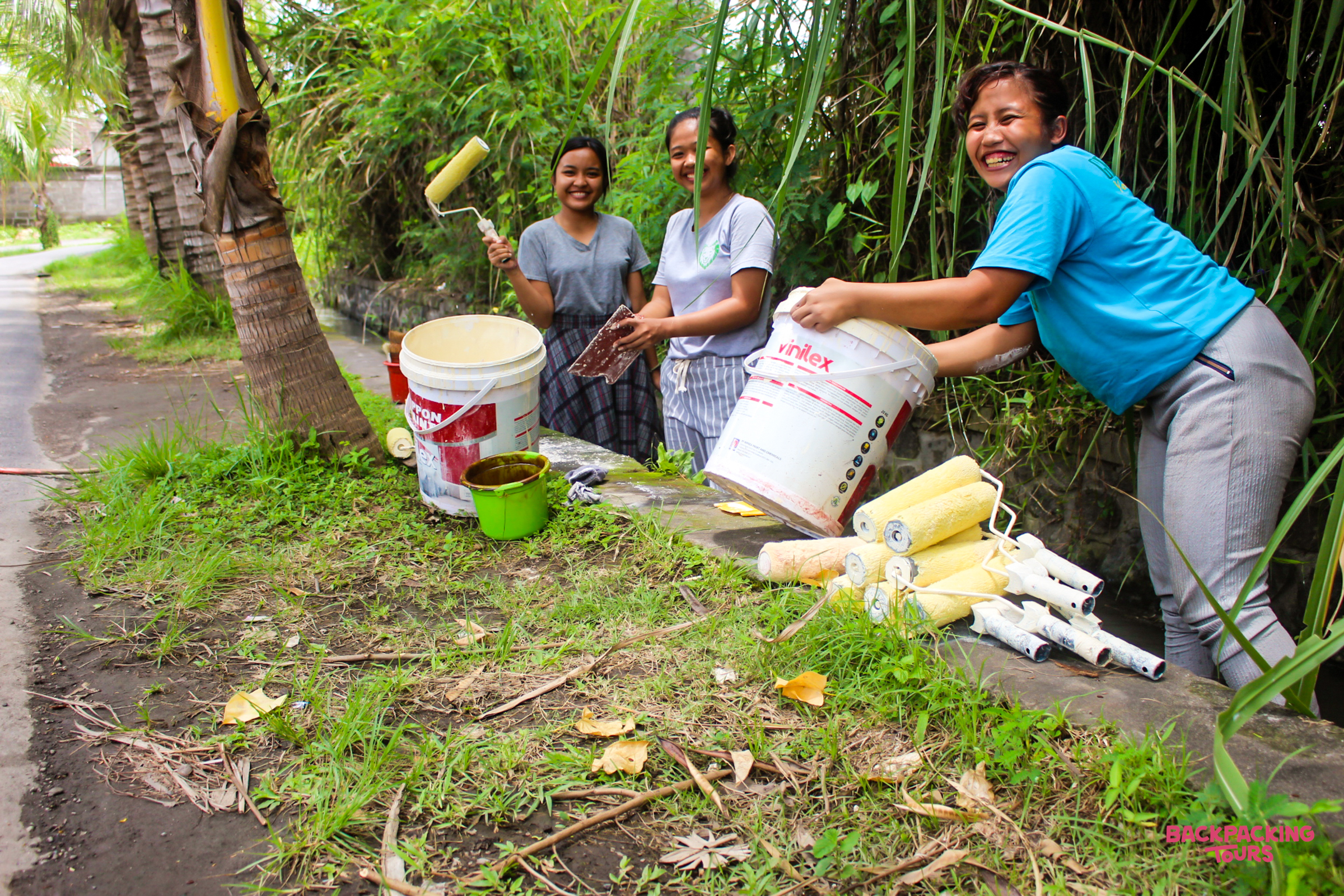 Three smiling women prepare to paint outdoors, with rollers and buckets on the grass.