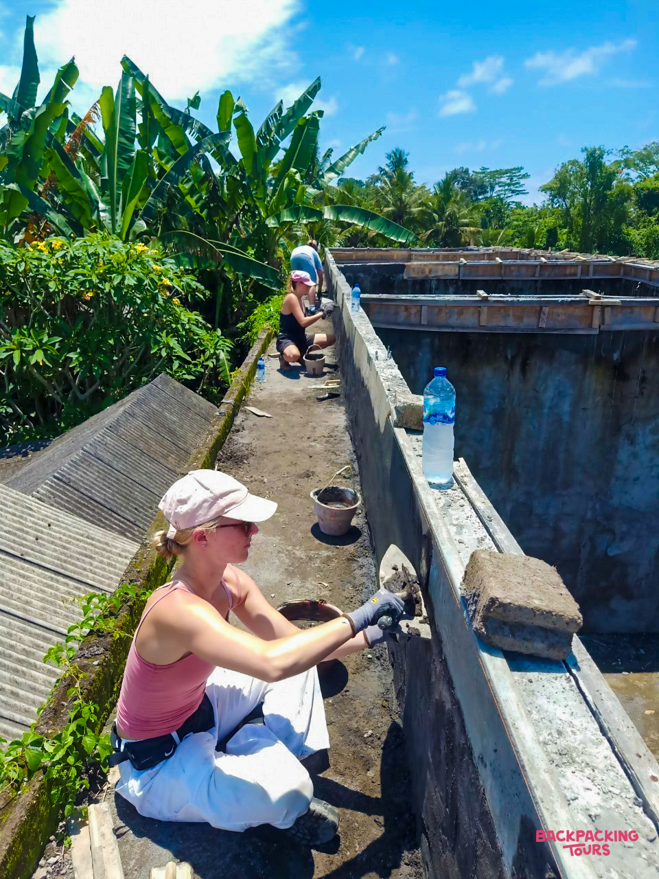 Volunteers work on a concrete structure under a bright blue sky surrounded by tropical plants in Bali.