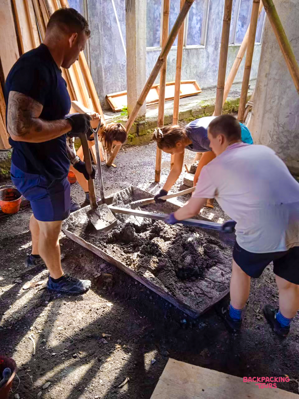 Volunteers enthusiastically mix cement by hand in a large tray at a construction site in Bali.