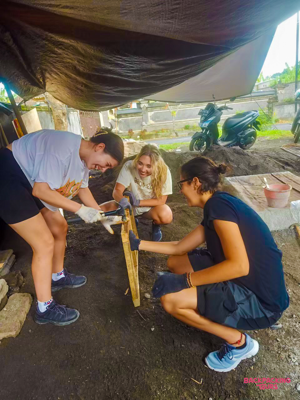 Three smiling women joyfully building a wooden structure together under a tarp in Bali.