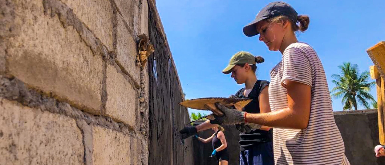 Two volunteers plastering a concrete block wall on a sunny day in Bali.