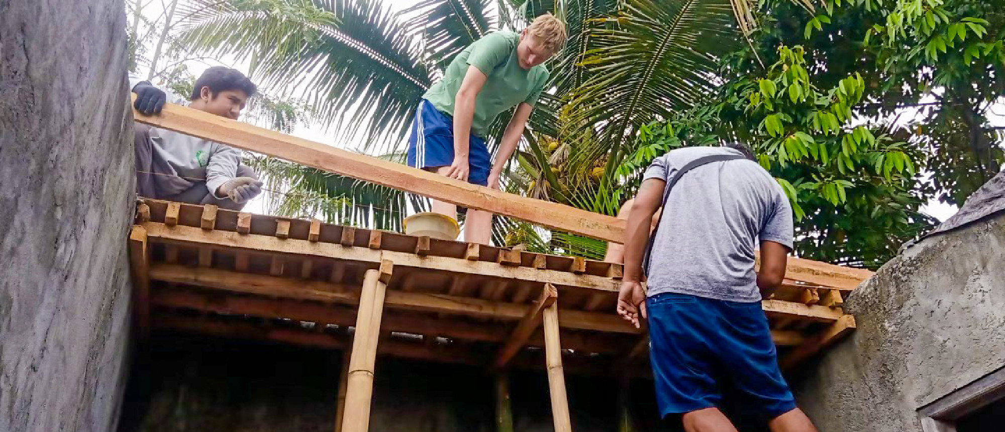 Three people work together to construct a wooden roof structure outdoors among tropical trees.