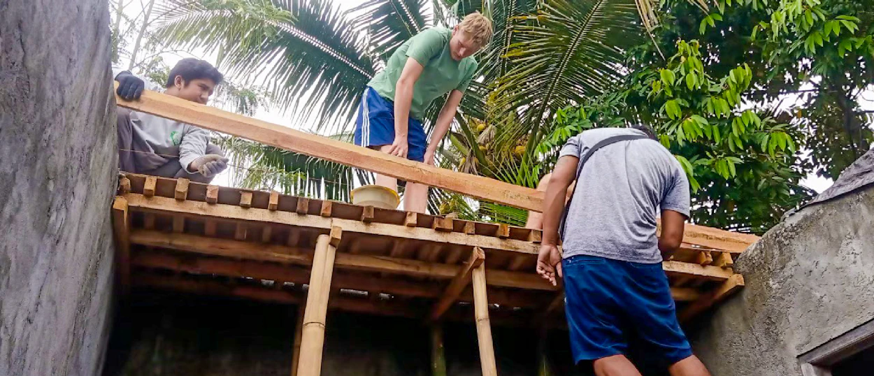 Three people work together to construct a wooden roof structure outdoors among tropical trees.