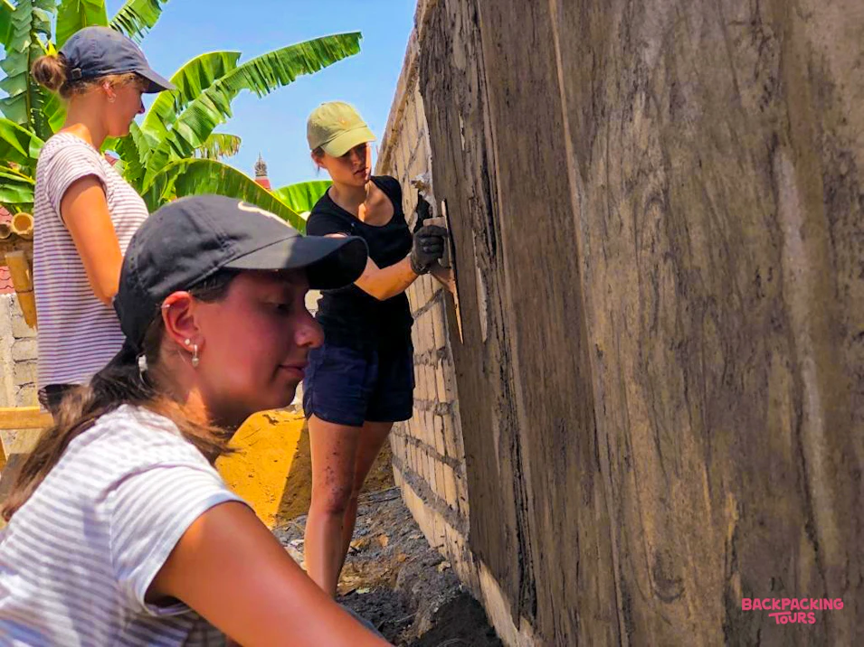 Three young women volunteers plastering a concrete wall at an outdoor construction project in Bali.