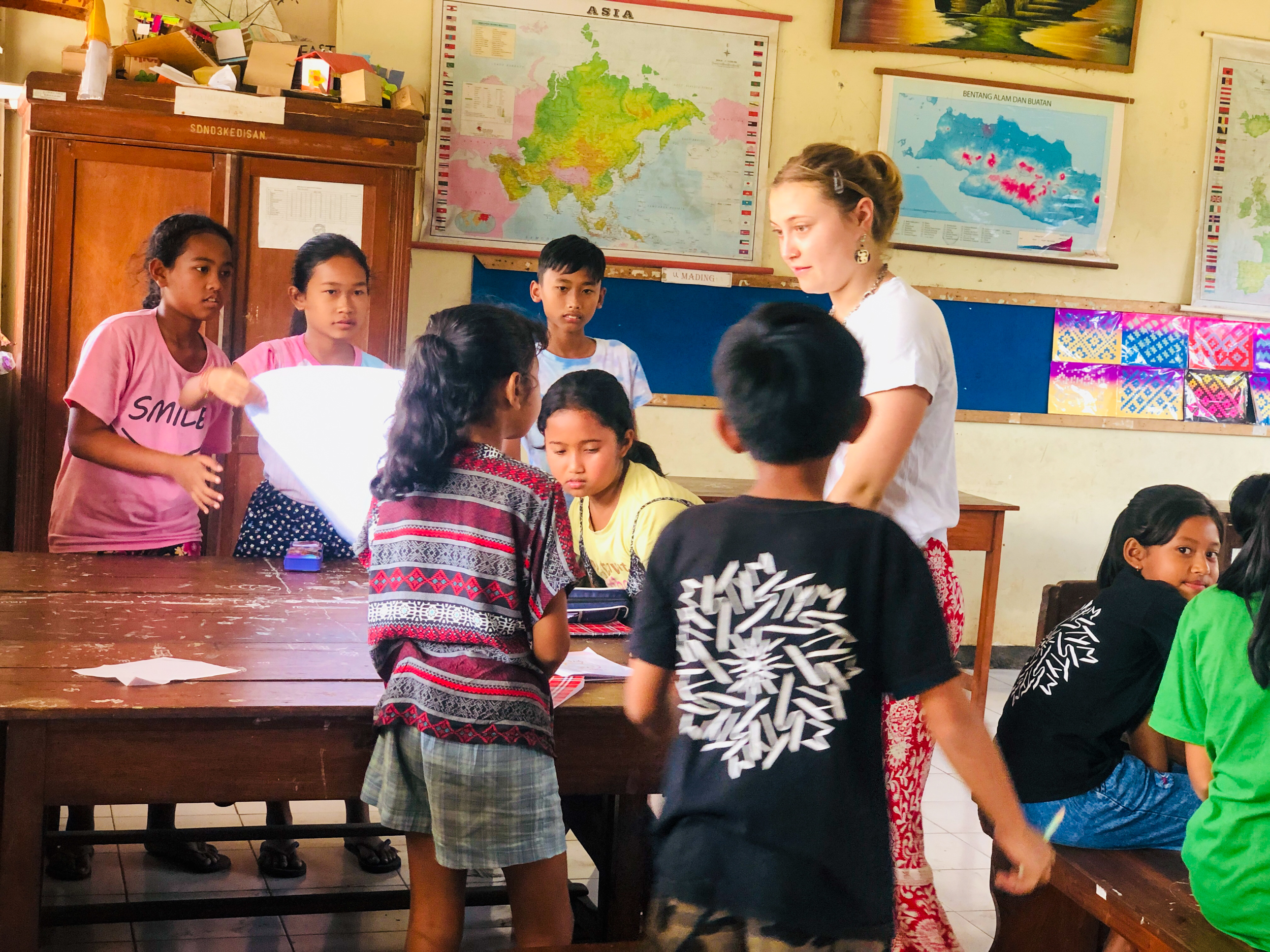 An adult volunteer interacts with children during a classroom activity, with maps on the wall, in Bali.
