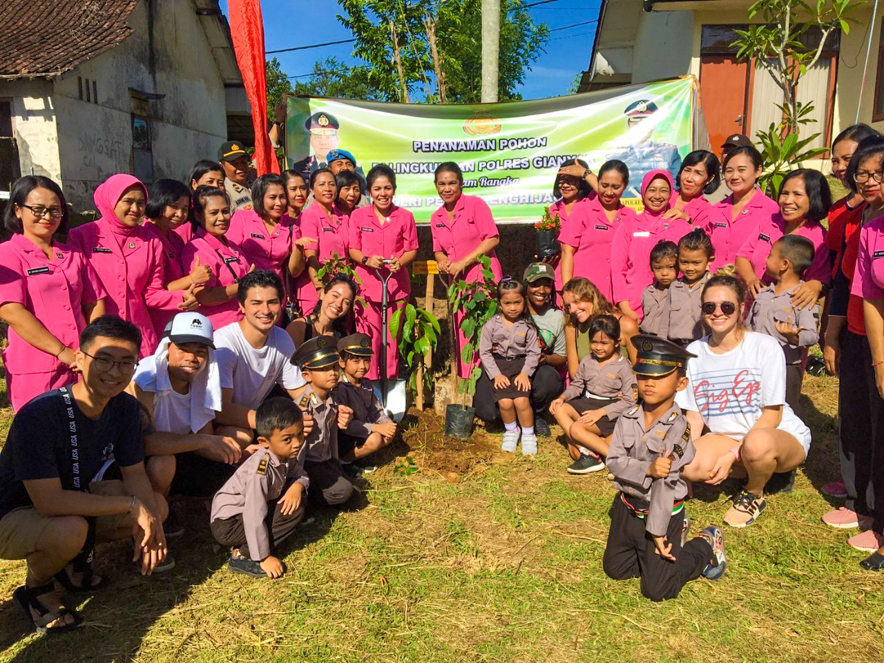 Smiling Balinese community members and children, some in uniform, gather around newly planted trees.