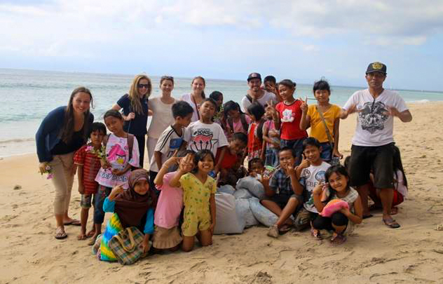 A large group of smiling children and adults pose together on a sandy beach in Bali.