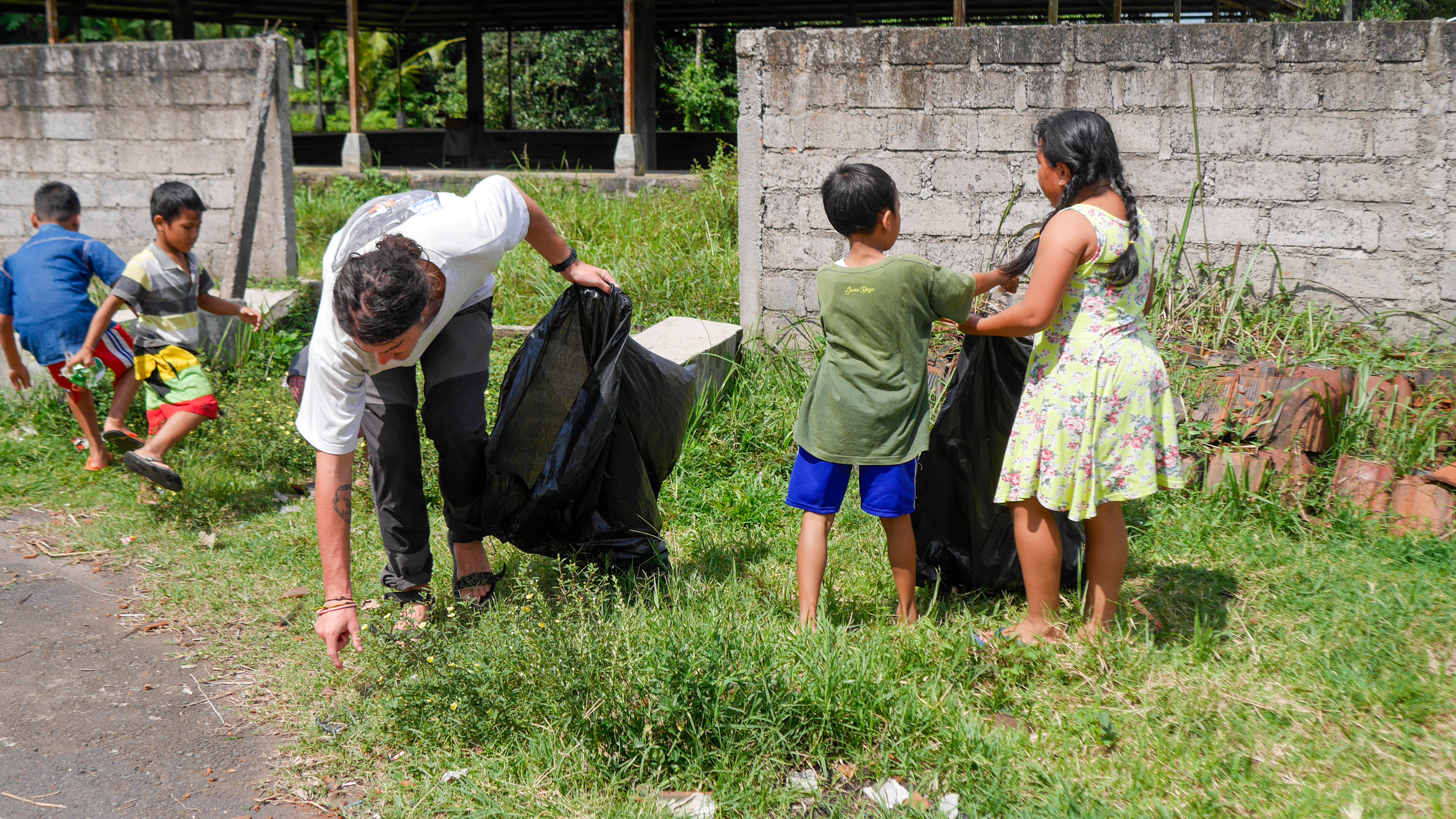 Volunteers, including children and an adult, diligently pick up litter from a grassy area in Bali.
