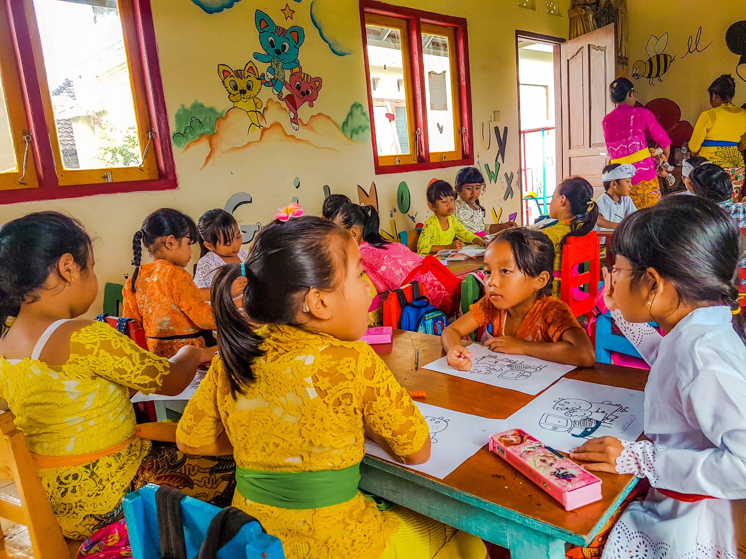 Young children in traditional Balinese clothing drawing and learning in a vibrant kindergarten classroom.