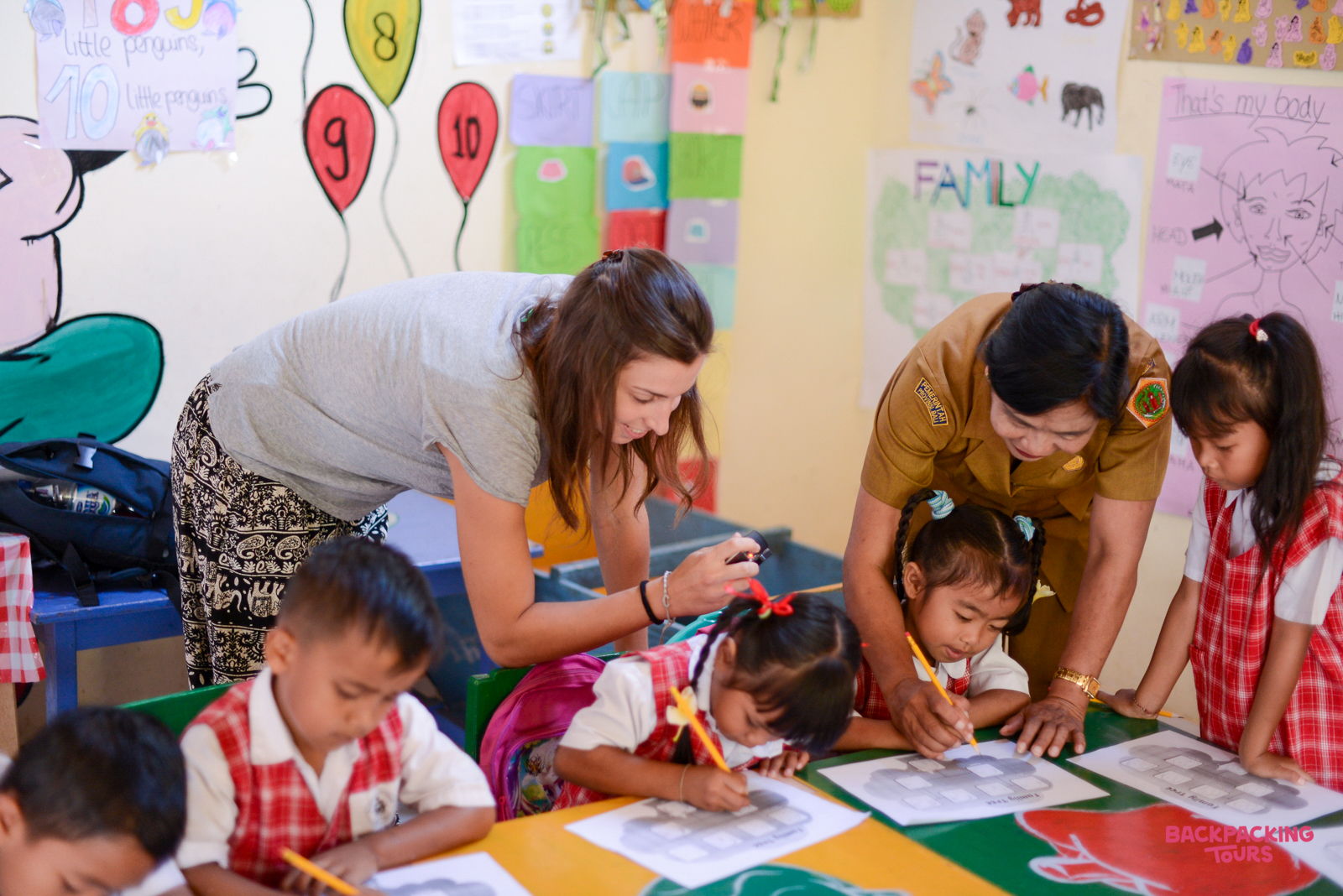 A volunteer and teacher help young children draw and learn in a colorful Bali kindergarten classroom.