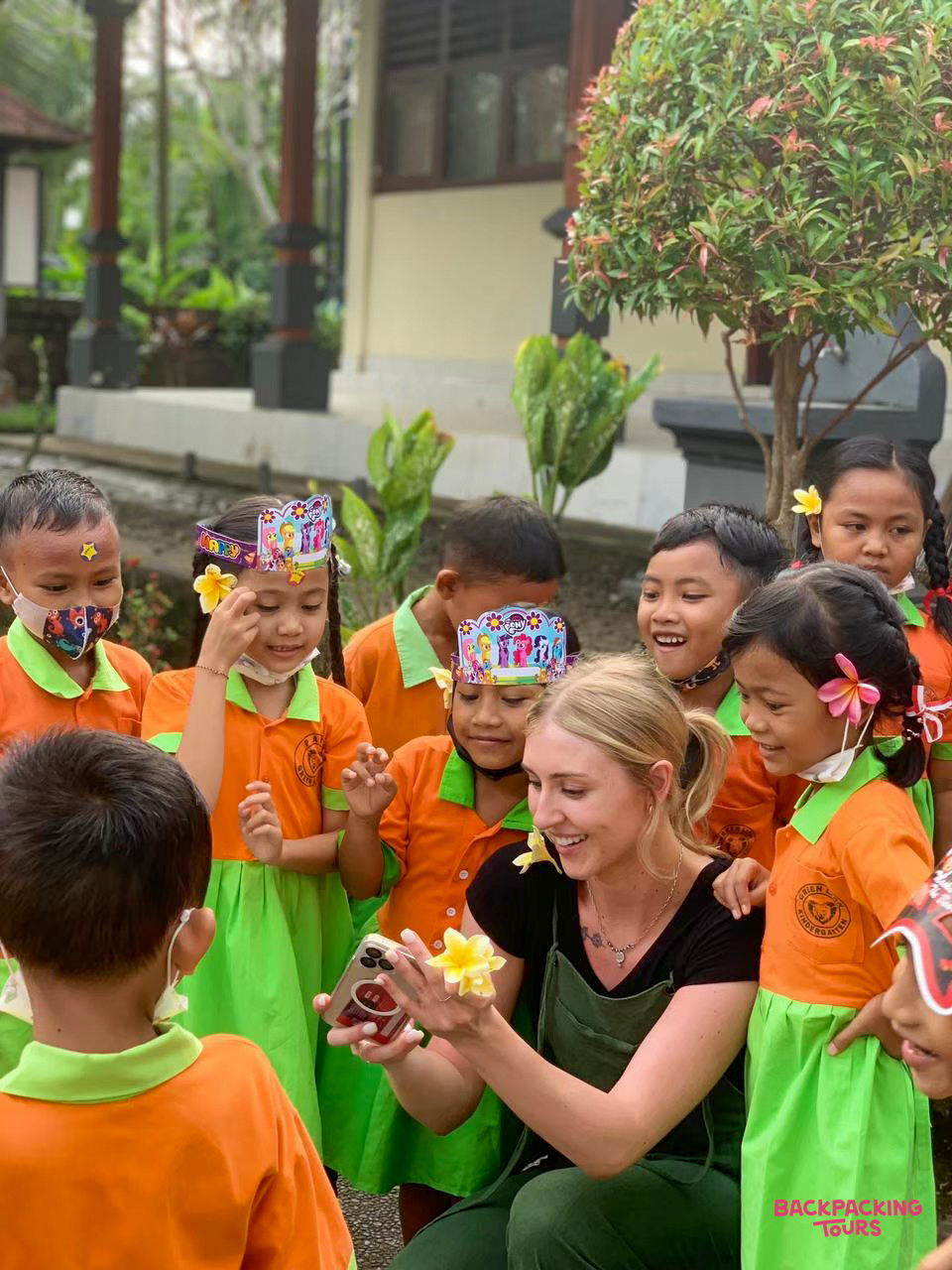 A smiling volunteer shares a moment with joyful Balinese kindergarten children, holding a phone and flower.
