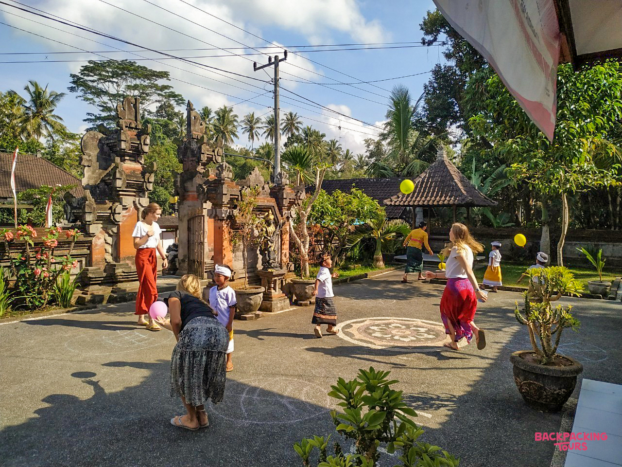 Happy children and volunteers play with colorful balloons in a sunny Balinese village courtyard.