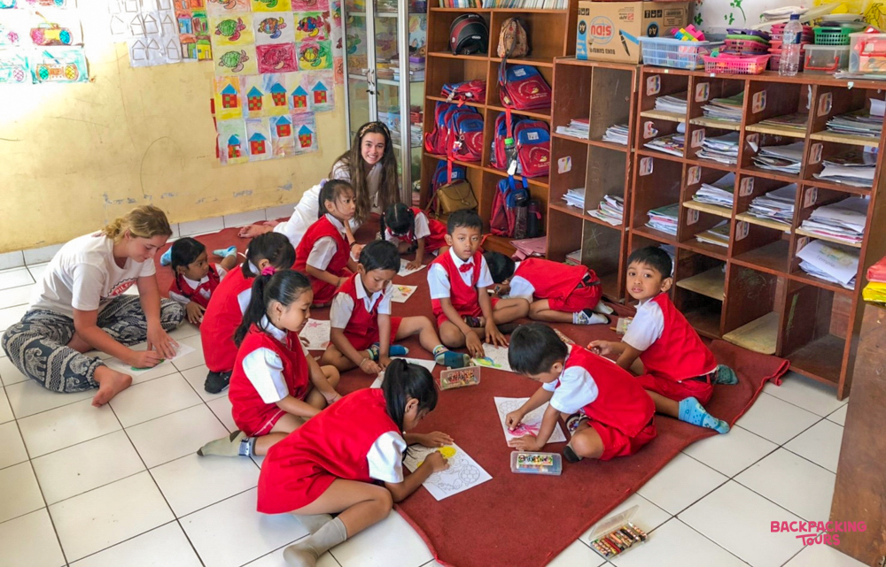Young children in red uniforms draw and learn with volunteers in a bright Bali classroom.