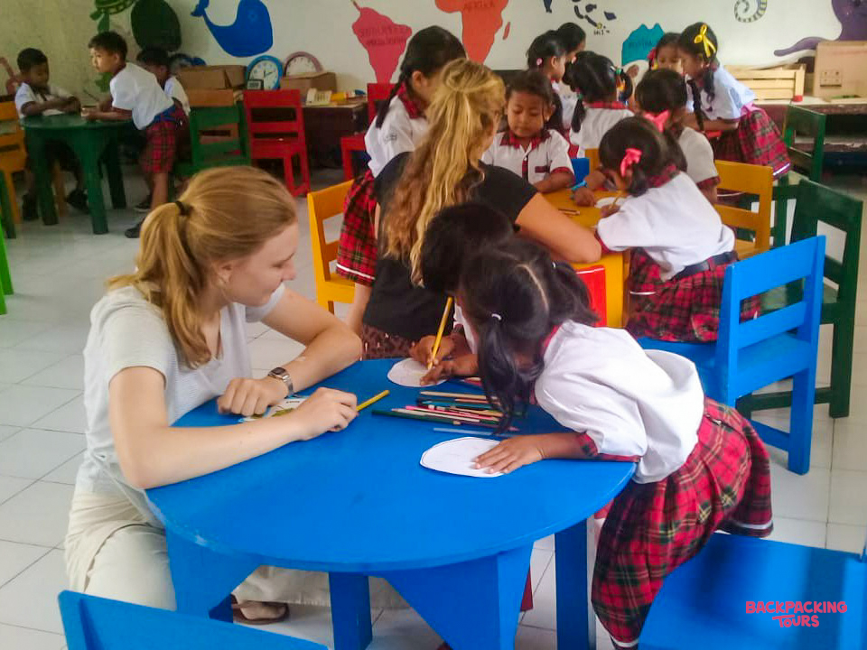 A volunteer helps a young girl draw at a blue table in a vibrant kindergarten classroom in Bali.