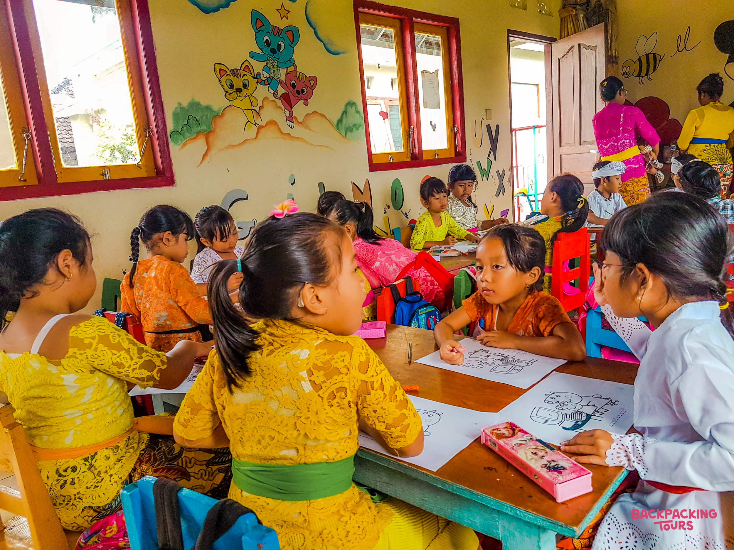 Young children in traditional Balinese clothing drawing and interacting at small tables in a vibrant kindergarten classroom.
