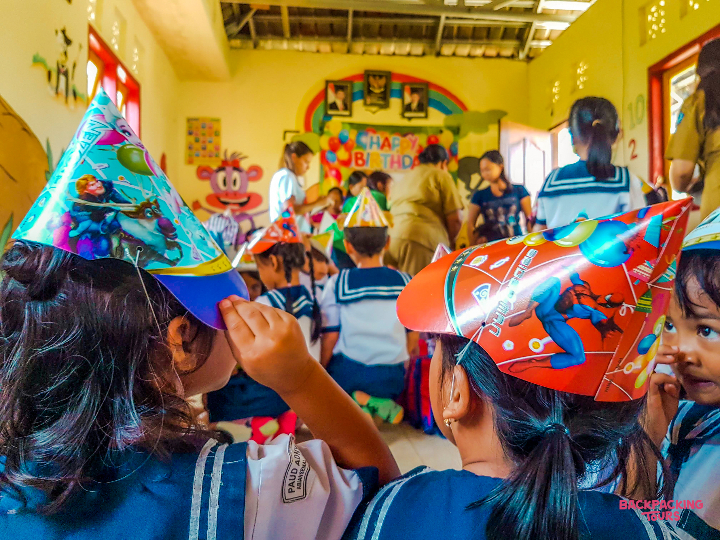 Children in party hats celebrate a birthday in a brightly decorated kindergarten classroom in Bali.