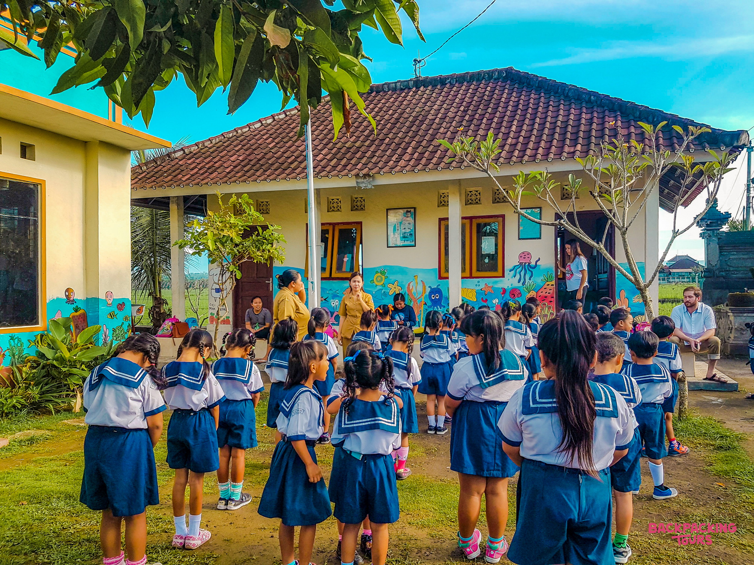Young children in blue and white uniforms gather outside a colorful kindergarten building in Bali.