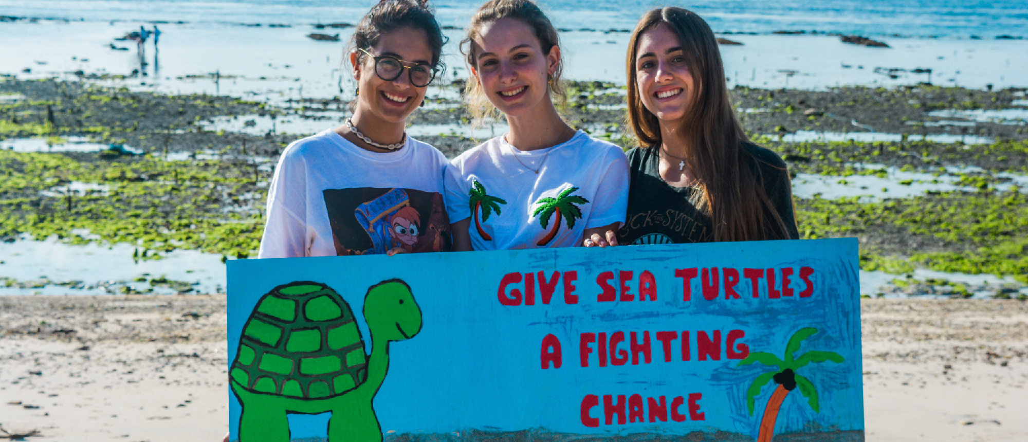 Smiling women hold a turtle conservation sign on a sandy beach with shallow water in Nusa Penida.