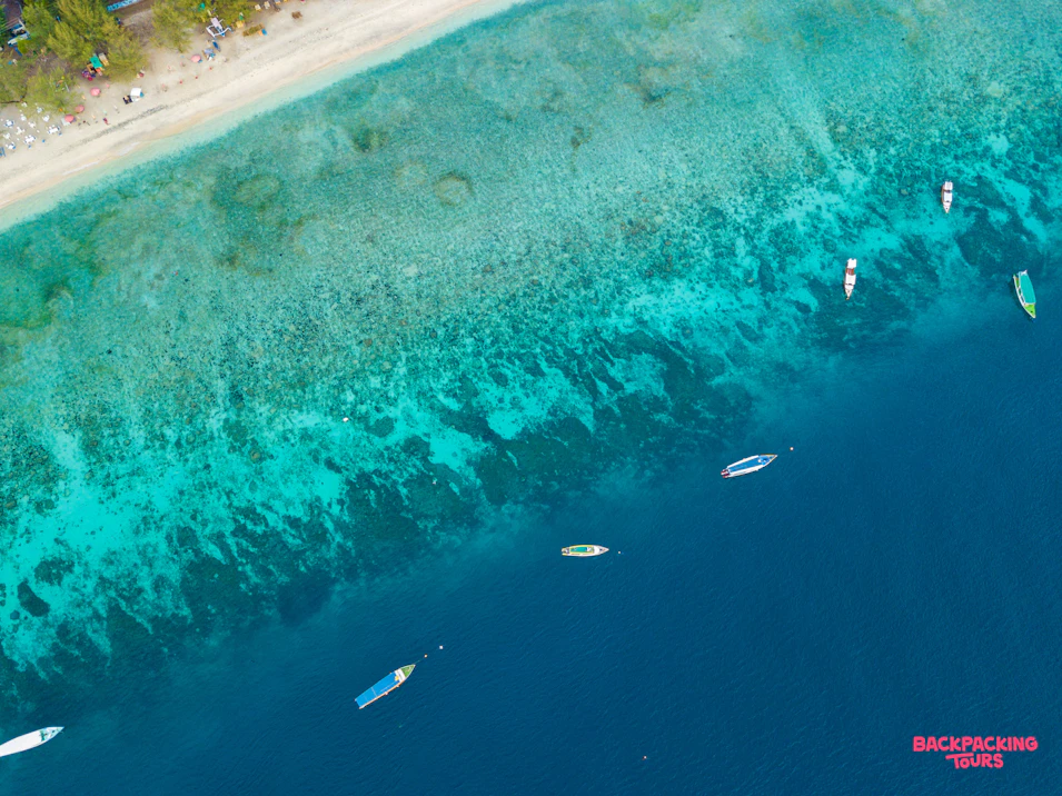 Boats dot the clear turquoise waters off a sandy Bali beach from an aerial perspective.