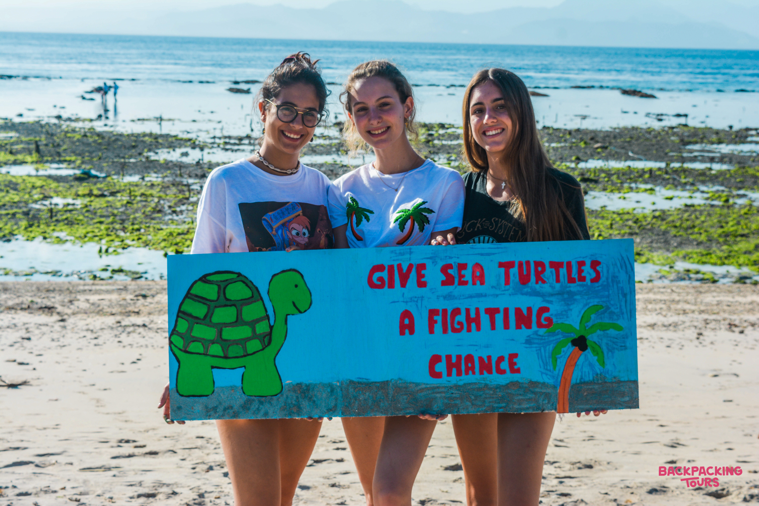 Three smiling women hold a sign for turtle conservation on a sunny beach in Nusa Penida.