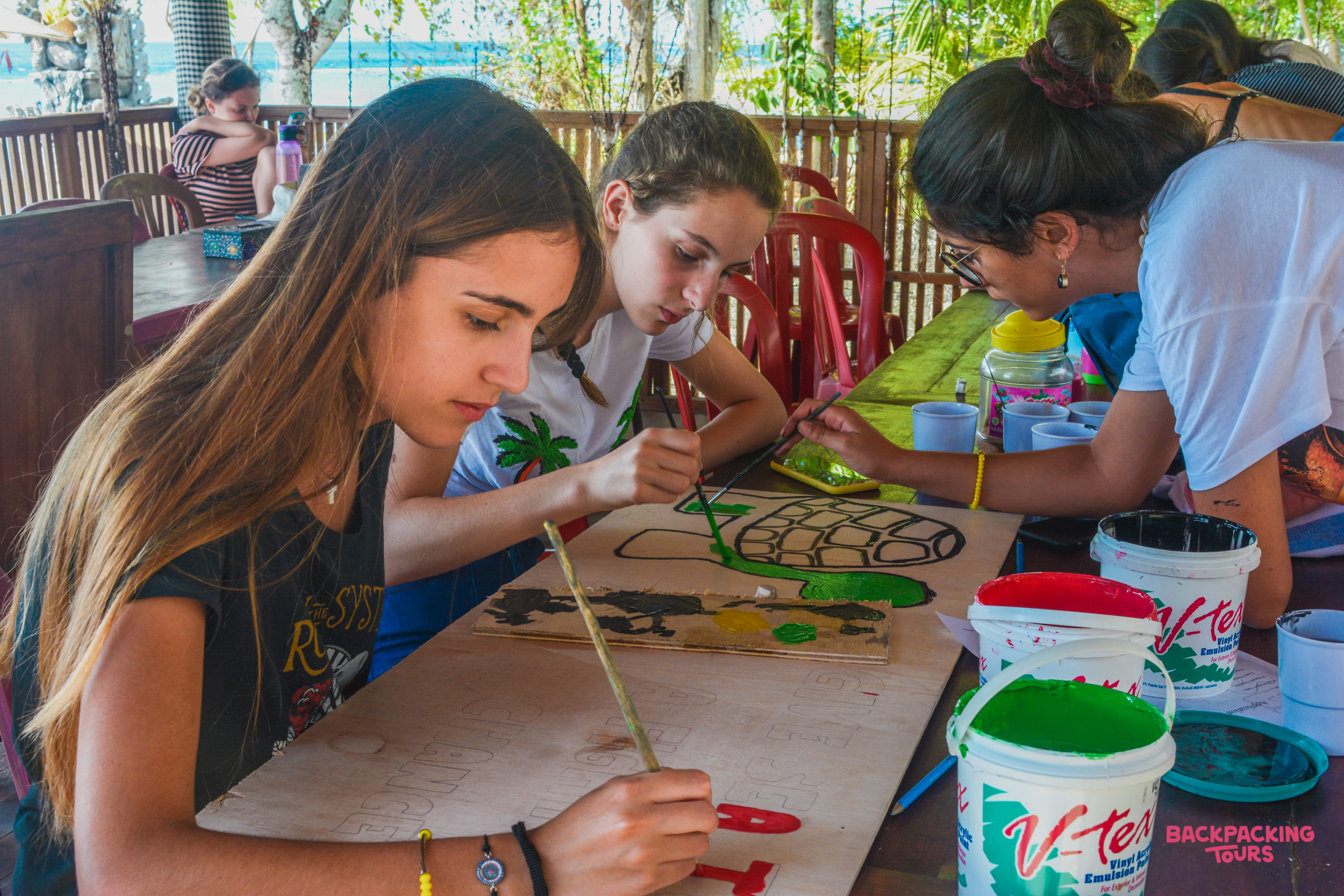 Three young women focused on painting turtle-themed designs on a wooden board outdoors in Nusa Penida.