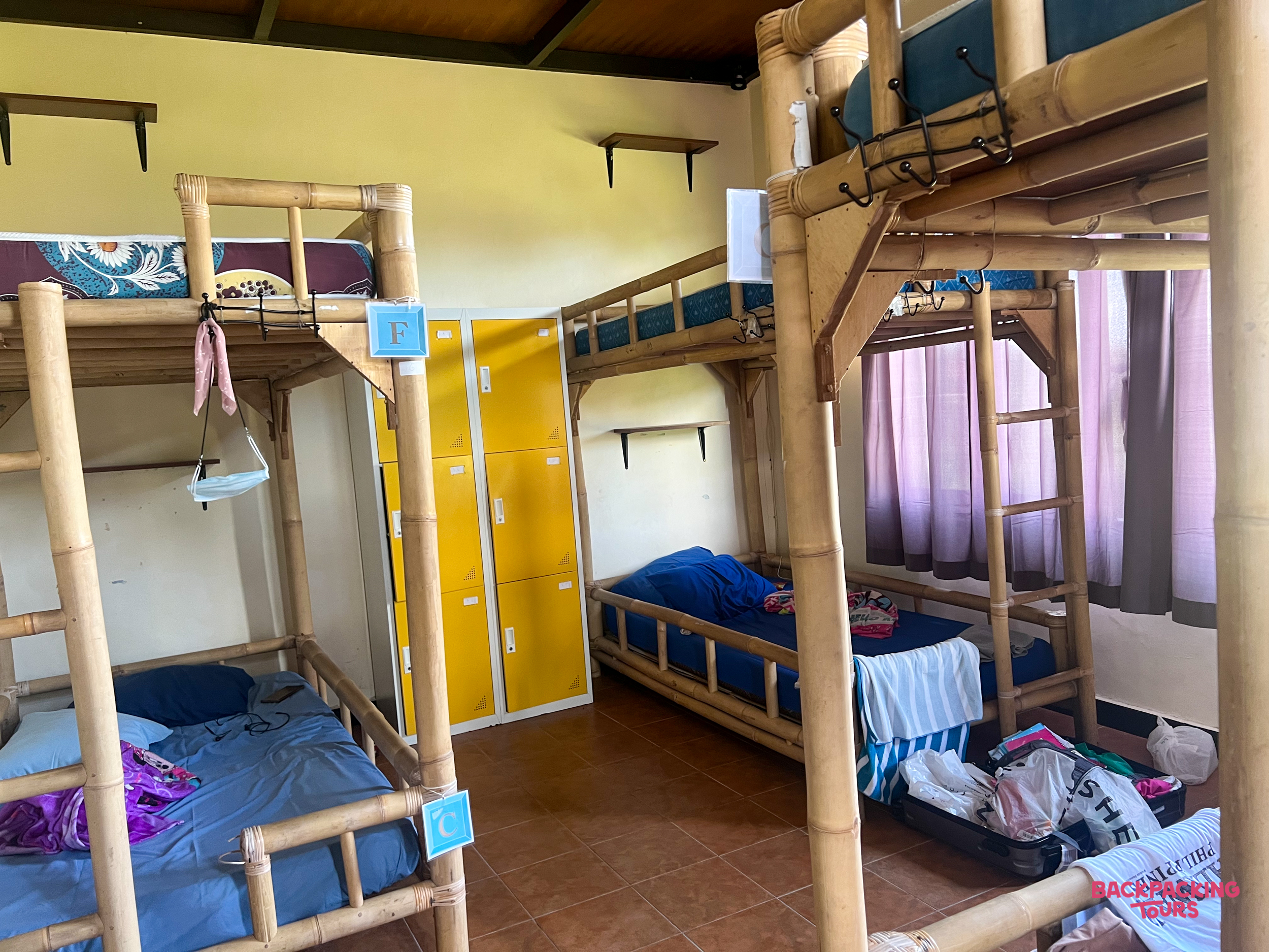 A bright hostel room featuring sturdy bamboo bunk beds and yellow lockers for guests' belongings.