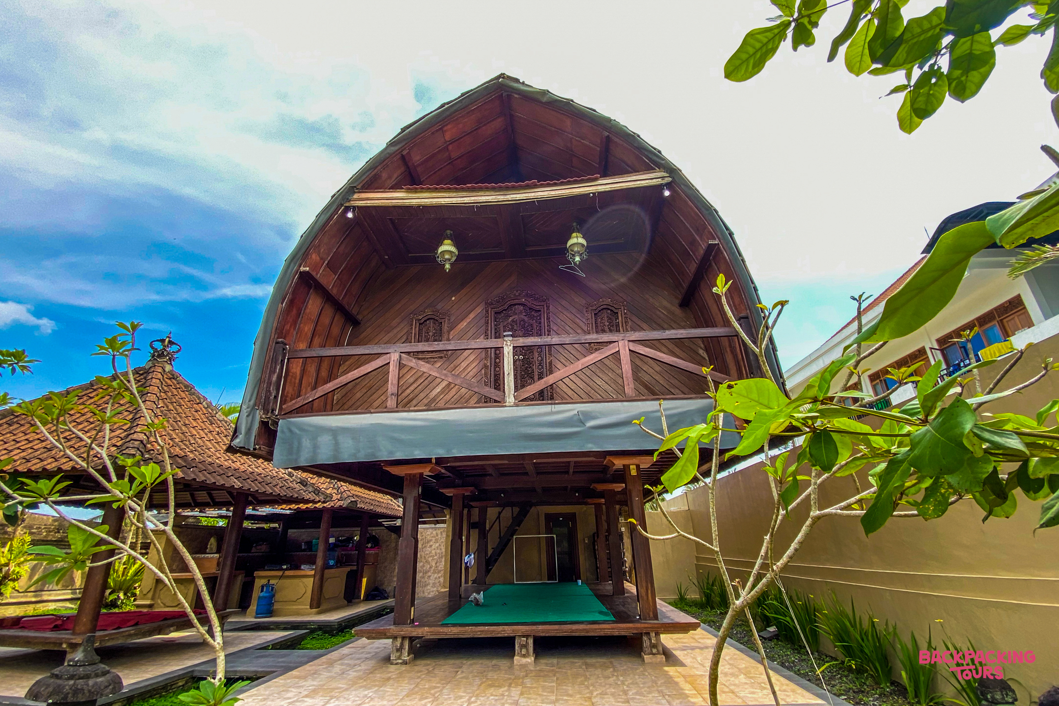 Traditional Balinese wooden accommodation with a curved roof and open-air lower level, surrounded by lush plants.