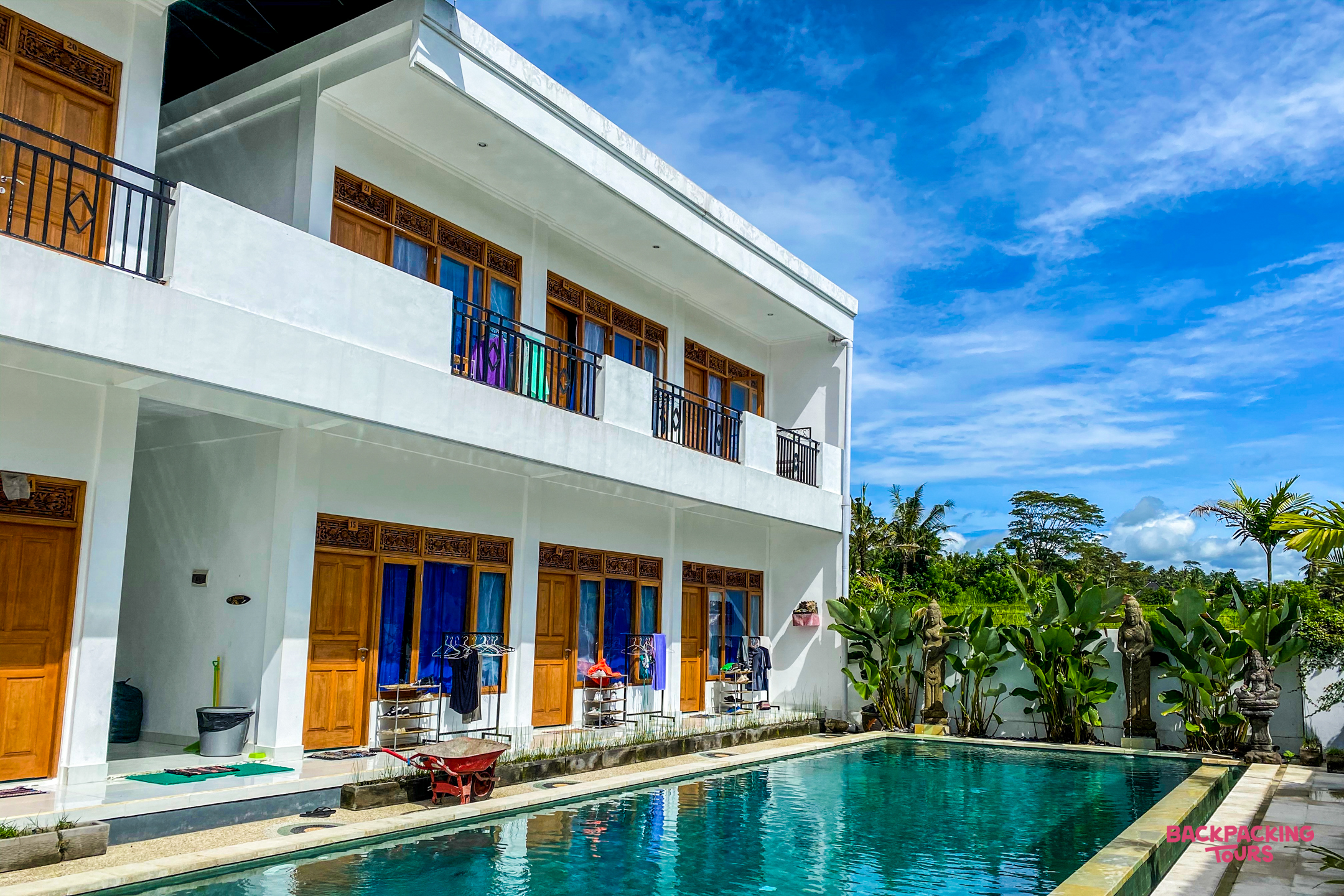 Bright white guesthouse with wooden doors and balconies overlooking a refreshing swimming pool in Bali.