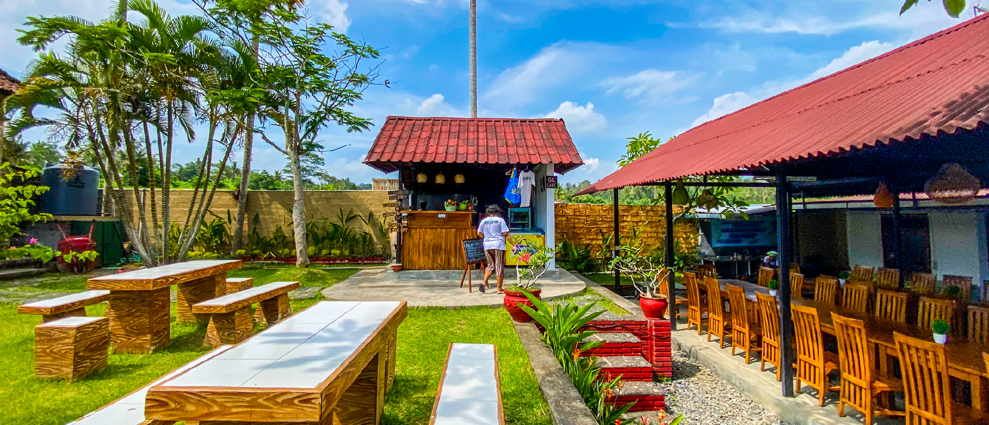 Sunny outdoor dining area with wooden tables, a small kiosk, and tropical plants in Bali.