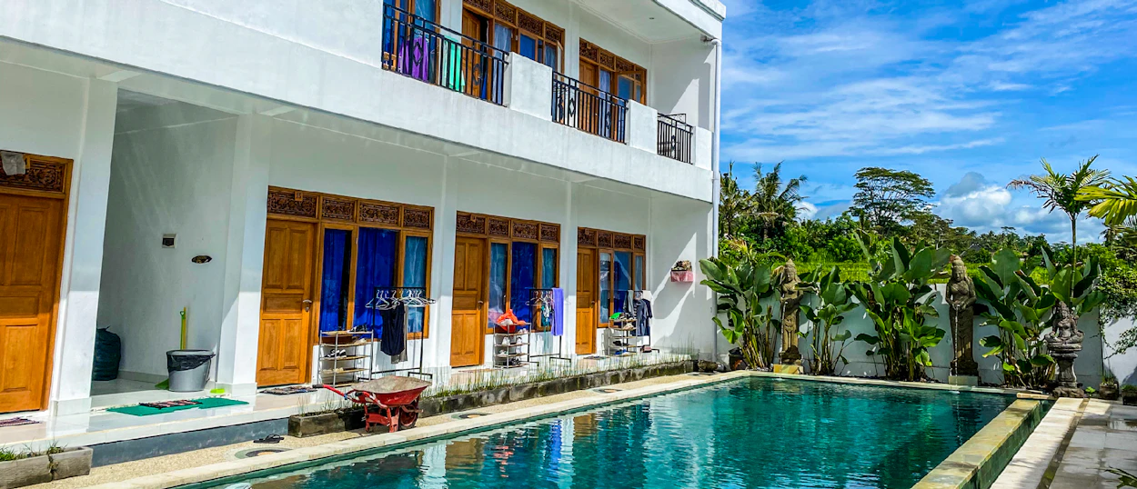 Modern white building with wooden doors and balconies next to a sparkling pool and tropical plants in Bali.
