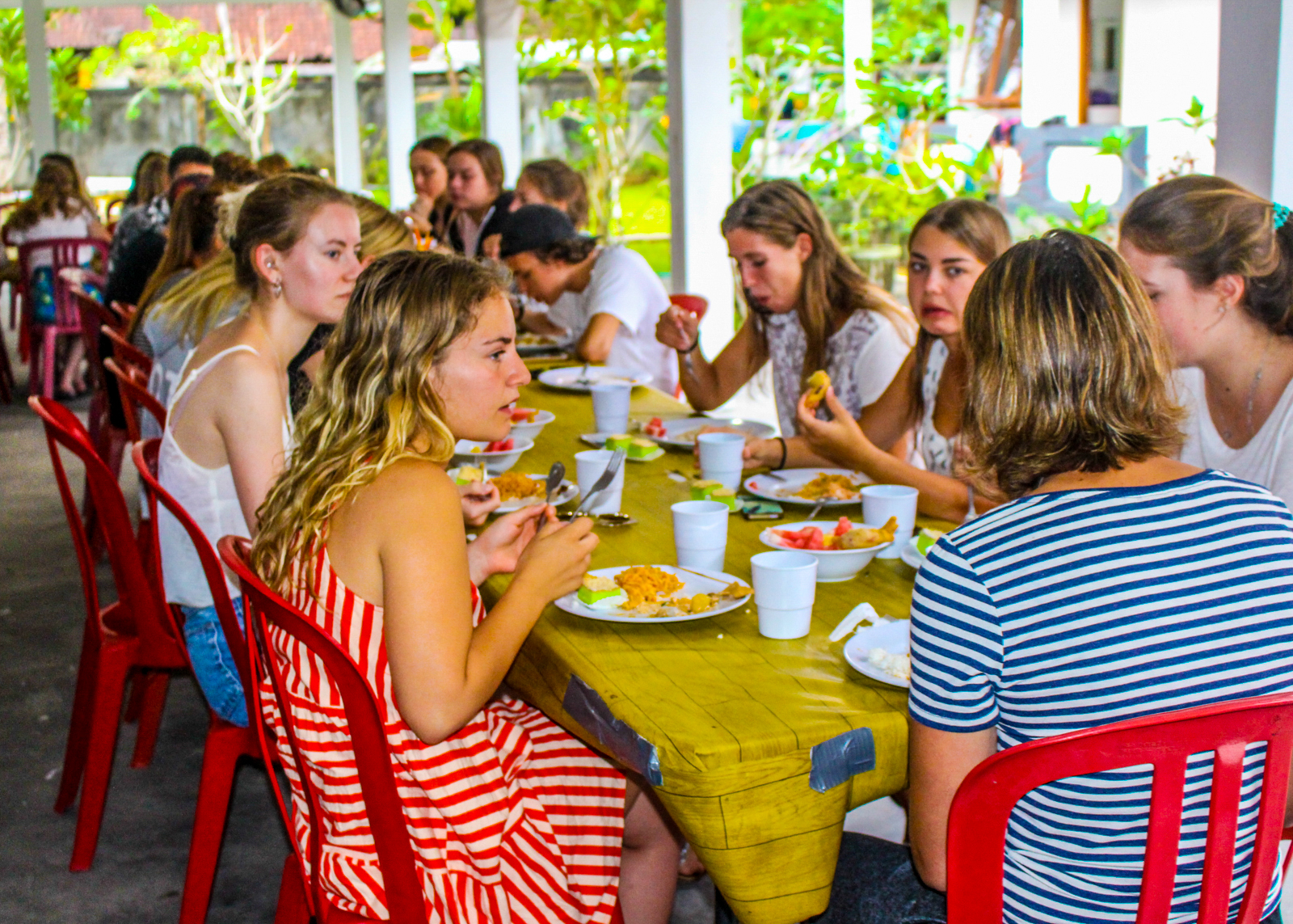 Young travelers share a casual meal at a long table in an open-air dining hall.