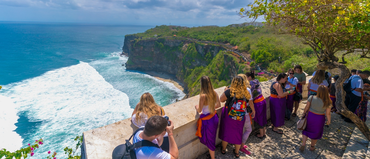 Visitors at Uluwatu temple overlook the vast ocean, crashing waves, and lush green cliffs of Bali.