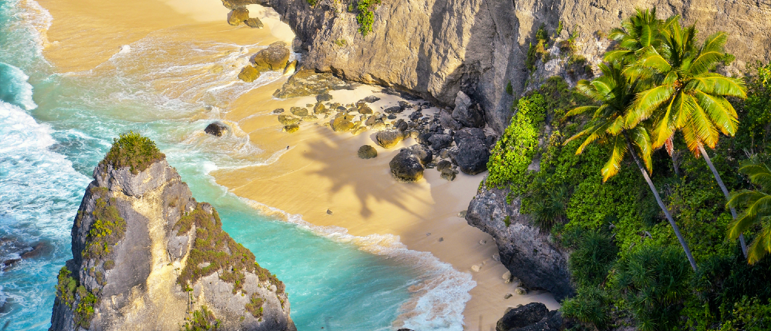 Stunning aerial view of a tropical beach in Bali with turquoise water, white sand, and towering cliffs.
