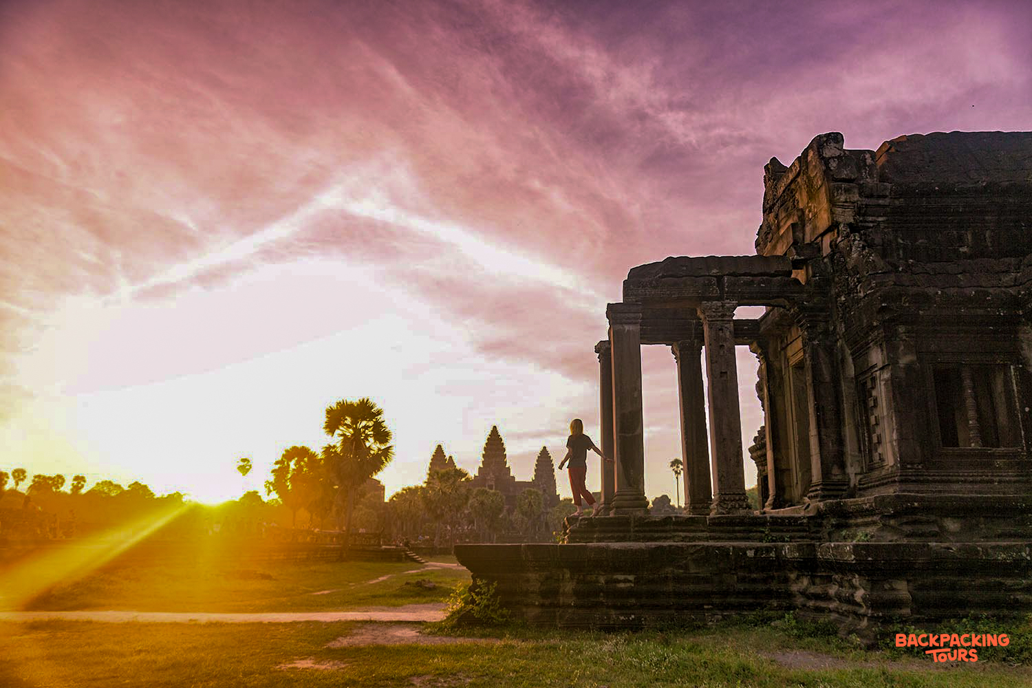 A traveler stands on ancient temple ruins, watching the vibrant sunrise over Angkor Wat in Cambodia.