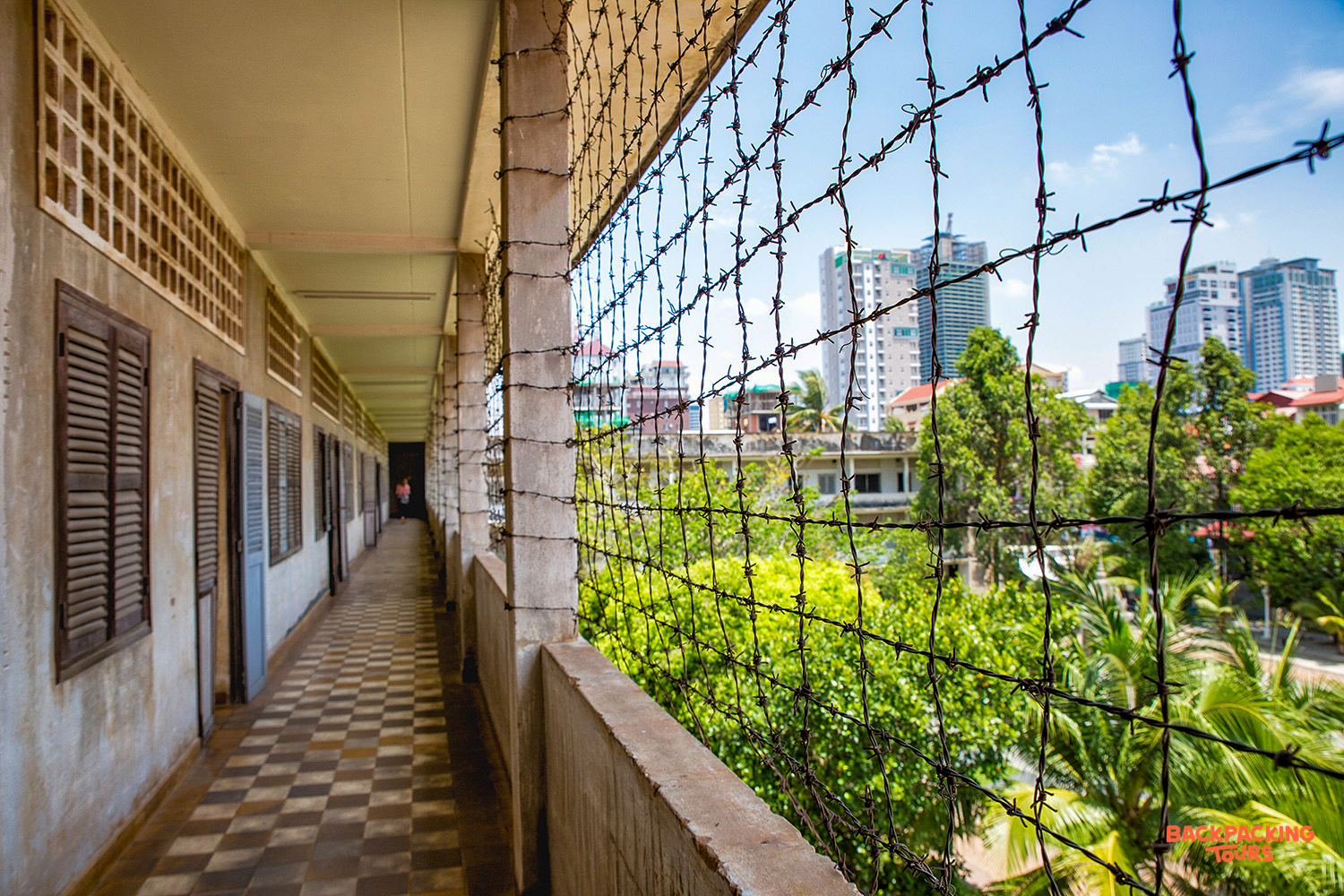 A long, somber corridor with barbed wire overlooking the modern Phnom Penh city skyline.