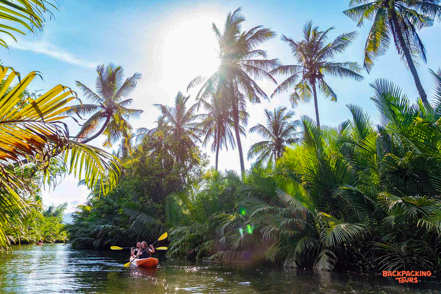 Two people kayak on a calm river, surrounded by lush tropical palm trees under a bright sun in Kampot.