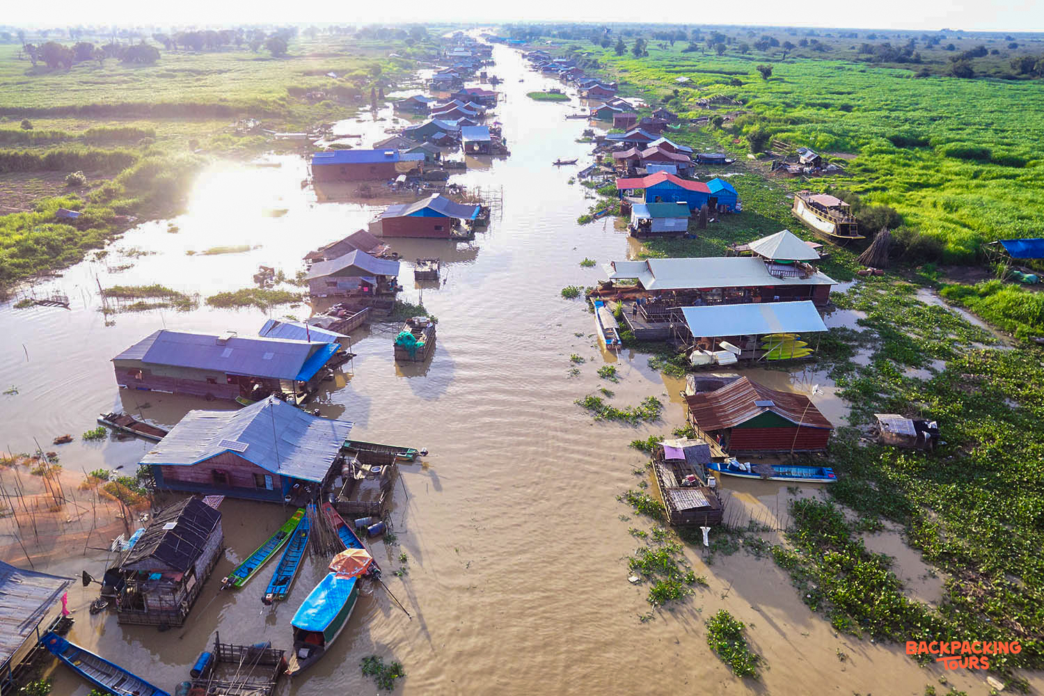 Peaceful floating village houses line a wide river surrounded by green fields in rural Cambodia.