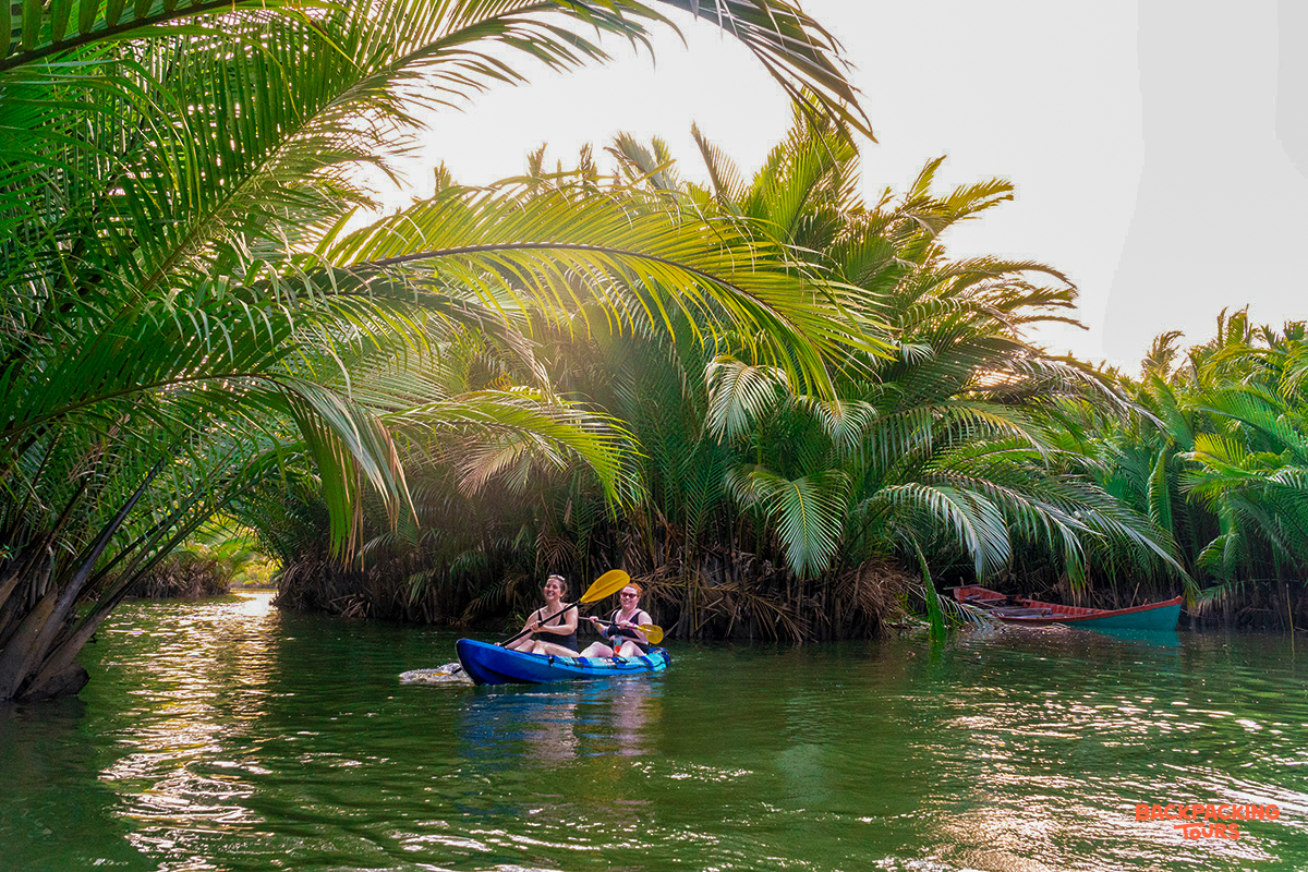 Two travelers peacefully kayak through a serene river surrounded by lush palm trees in Kampot.