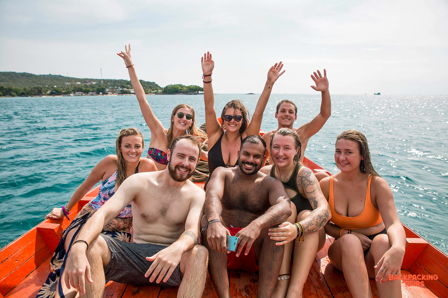Eight happy backpackers wave from a longtail boat on the clear blue waters of Koh Rong.