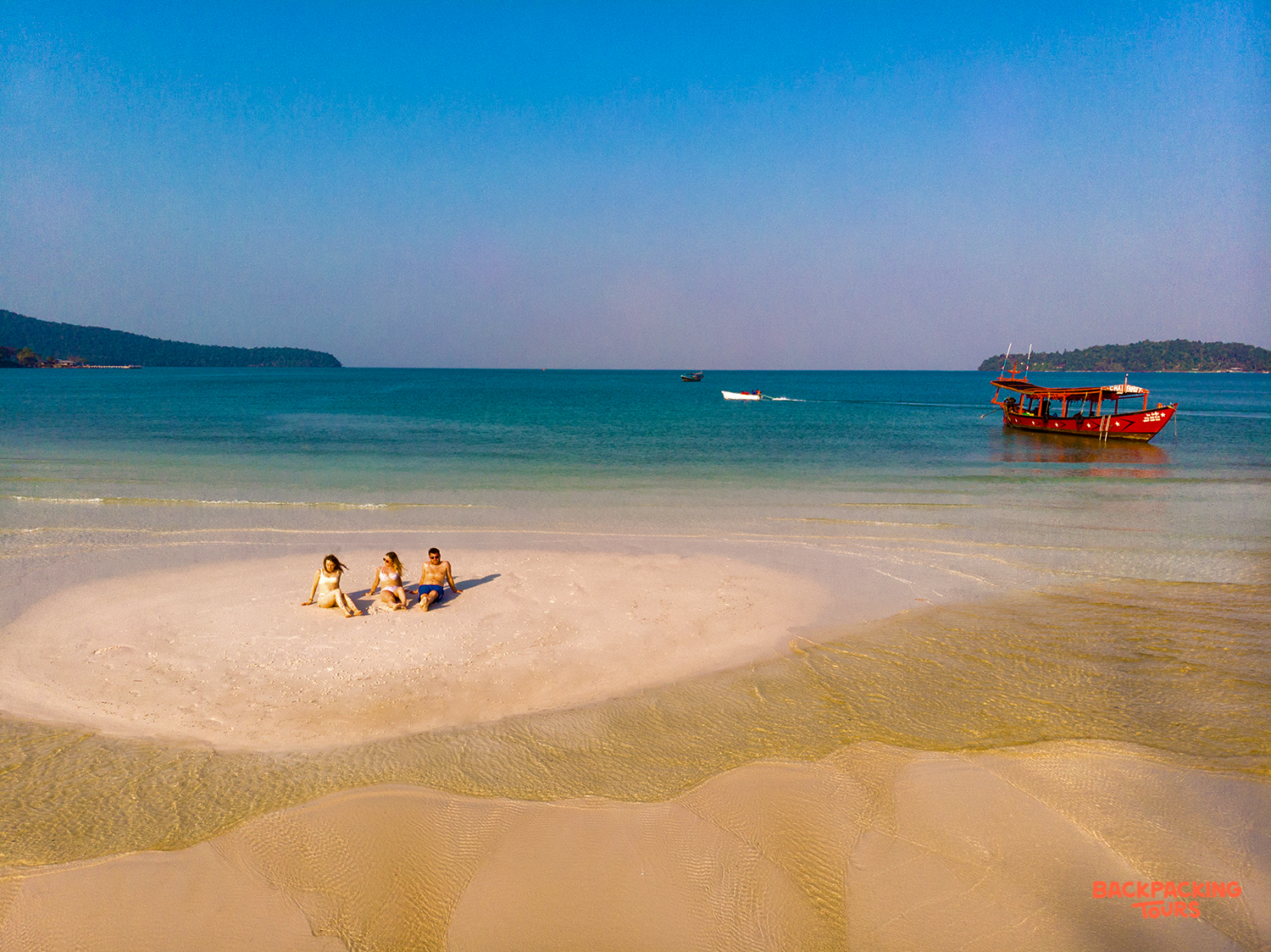 Three friends relax on a pristine sandbar surrounded by turquoise waters off Koh Rong, Cambodia.