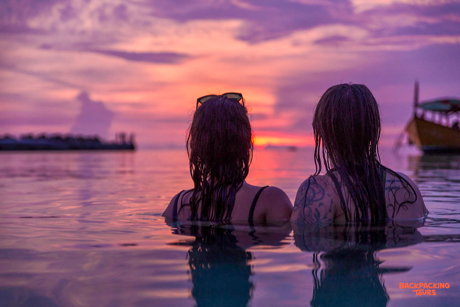 Two women relax in the ocean, watching a vibrant sunset over Koh Rong.